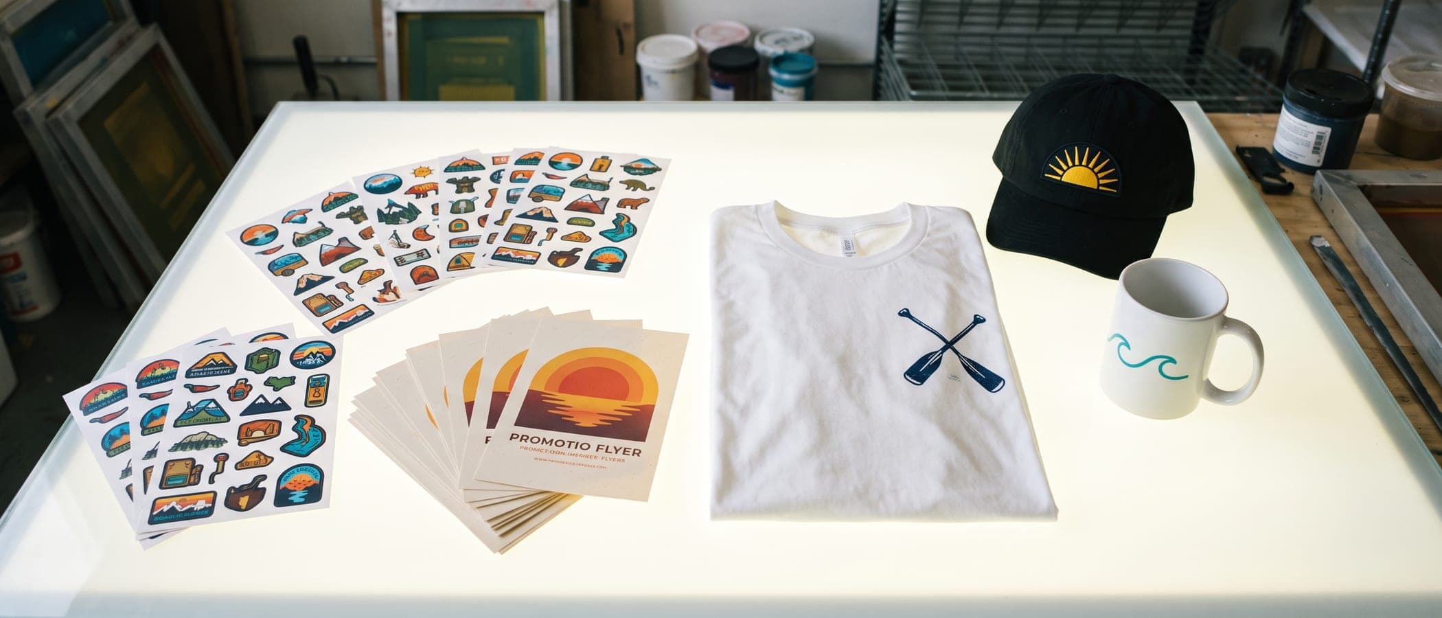 Produits imprimés disposés à plat sur une table lumineuse : textile, casquette, mug, stickers et flyers.